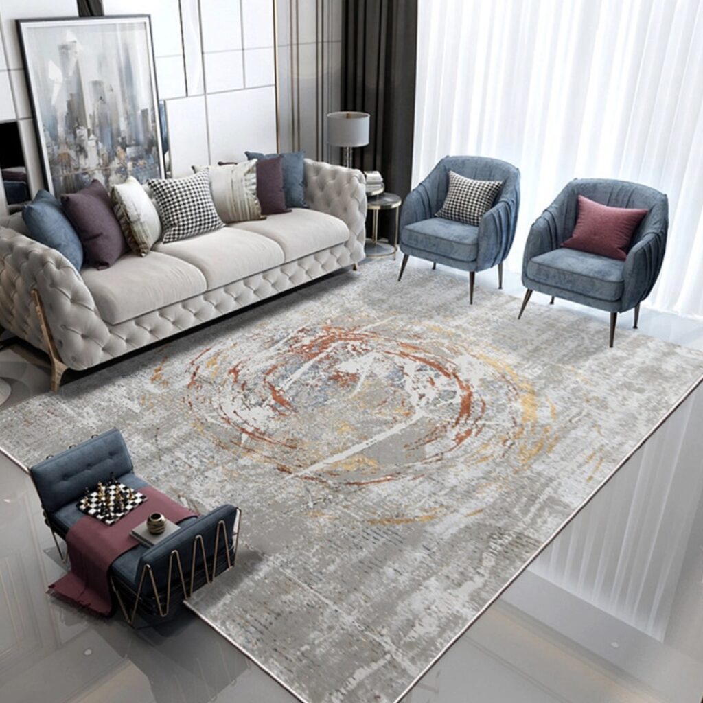 A modern living room featuring a contemporary abstract-patterned carpet in shades of grey, orange, and yellow. The room is furnished with a tufted beige sofa, two blue accent chairs, and decorative cushions, creating a chic and stylish atmosphere