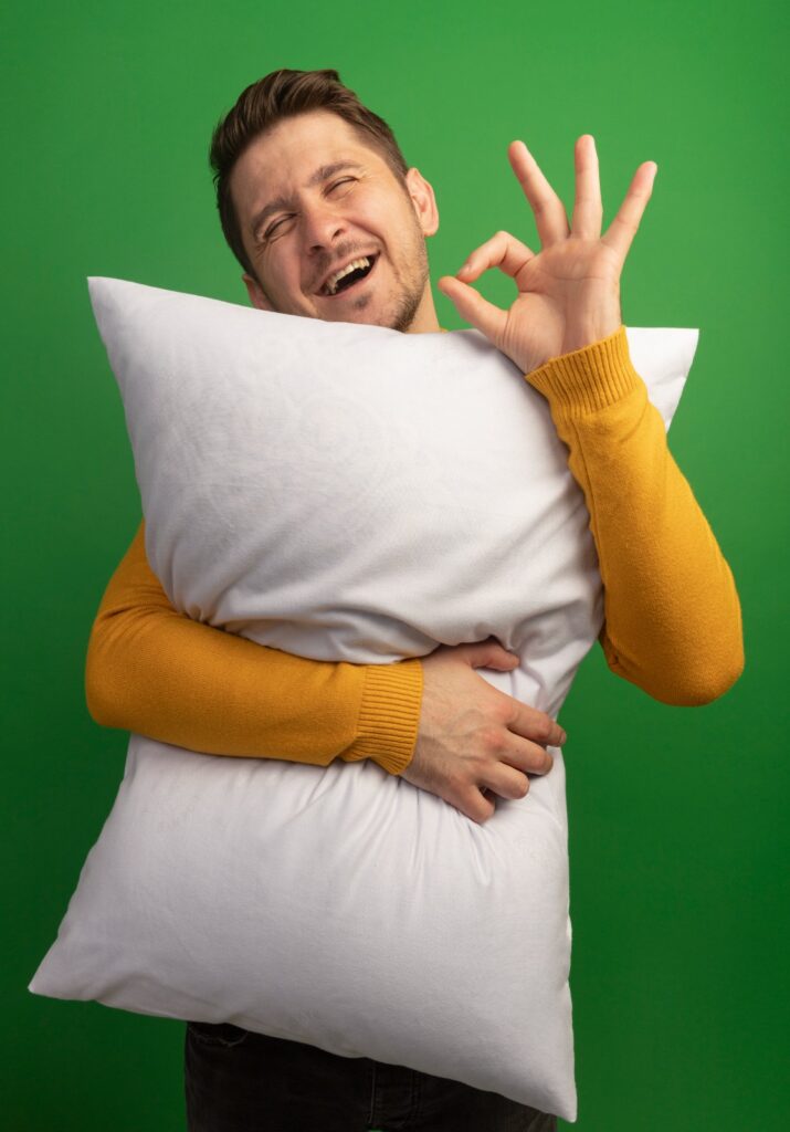 A young man holding a bed pillow make sign with his hand saying its perfect